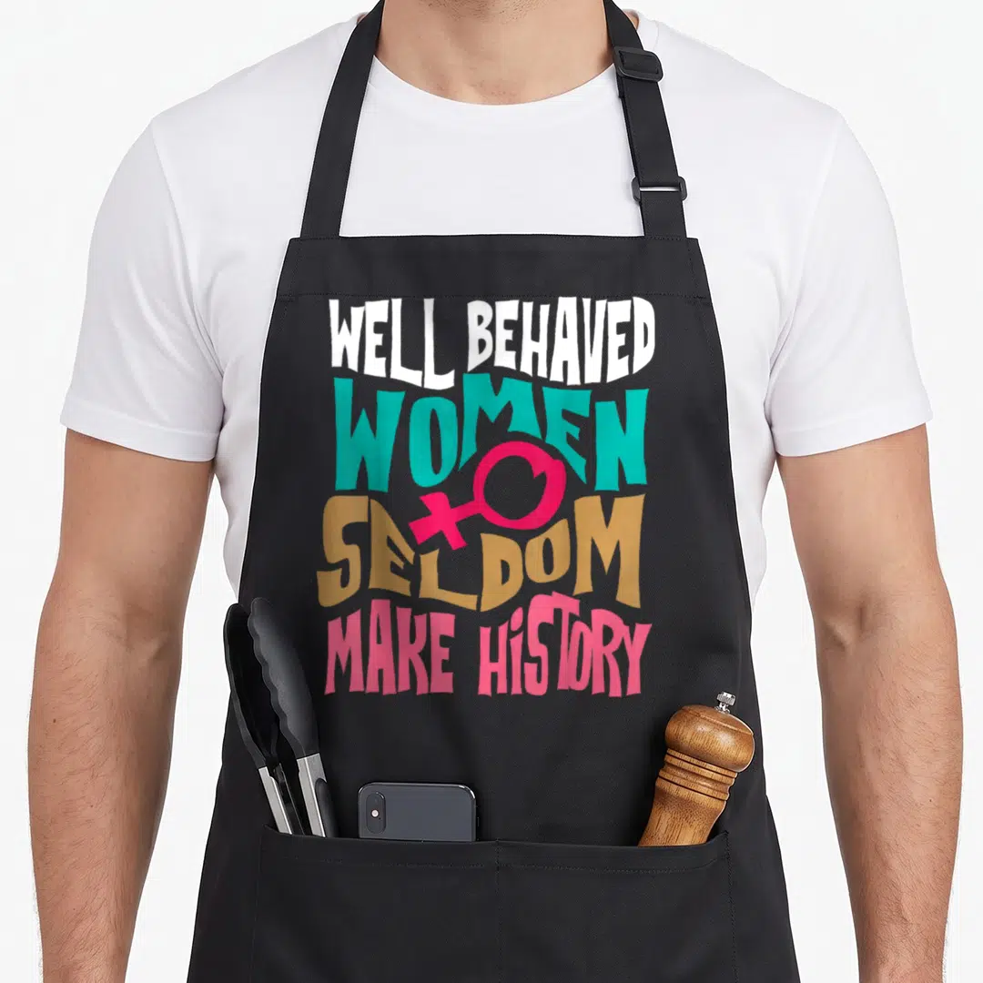 Behaved novelty apron - Stacked text reading WELL BEHAVED WOMEN SELDOM MAKE HISTORY with pink female symbol in white — shown on a black cotton canvas apron against a white background