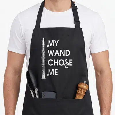 Wand clarinet apron - Vertical white clarinet next to stacked text MY WAND CHOSE ME with treble clef detail — shown on a black cotton canvas apron against a white background