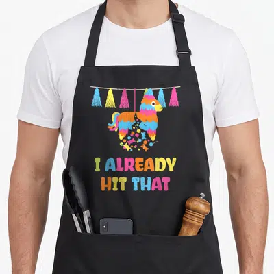 Vibrant mets apron - Cartoon piñata with candy and I Already Hit That text in rainbow colors — shown on a black cotton canvas apron against a white background
