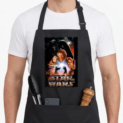Revenge droewors apron - Star Wars photo montage with Vader helmet and orange logo text — shown on a black cotton canvas apron against a white background