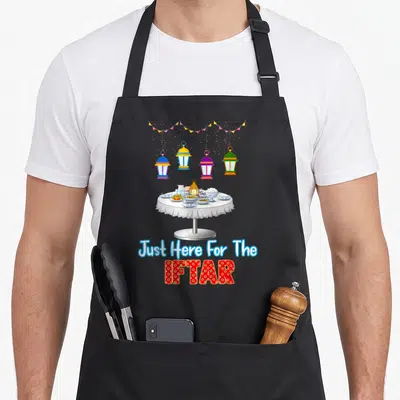 Ramadan table dad apron - Cartoon iftar table with hanging lanterns and Just Here For The IFTAR text — shown on a black cotton canvas apron against a white background
