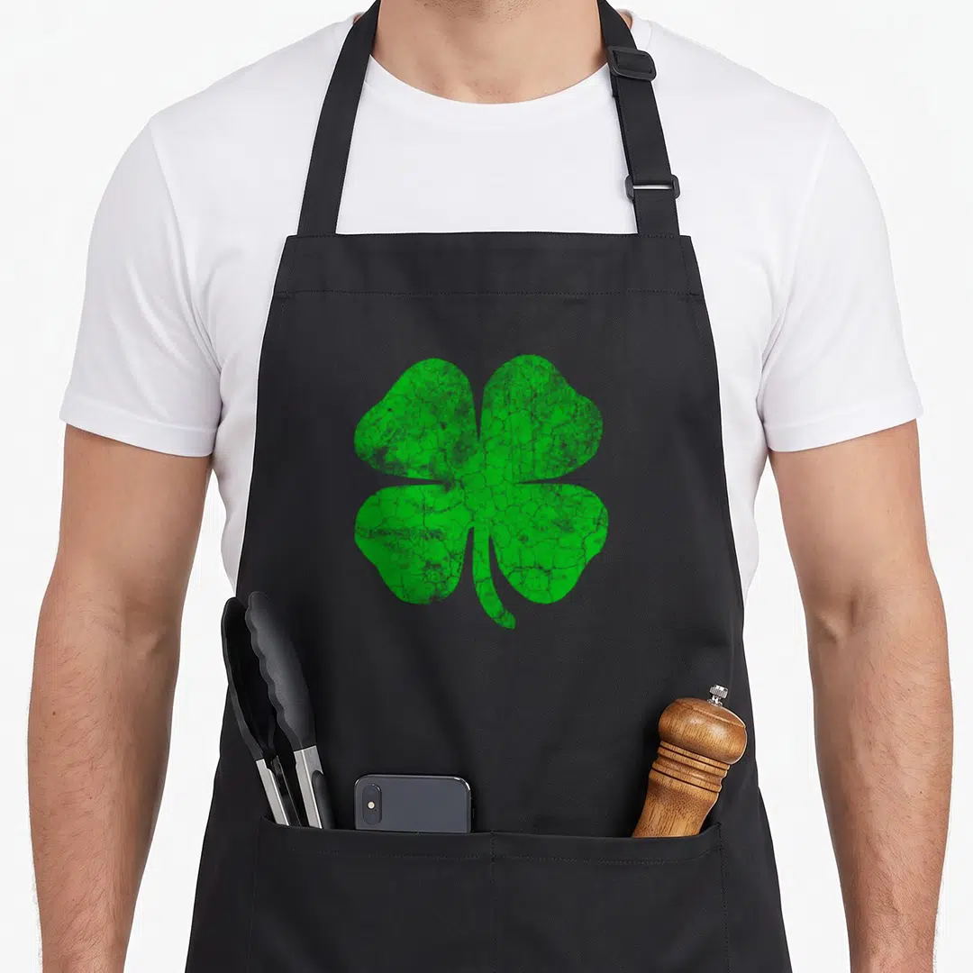 Irish clover st patrick day aprons - Distressed green four leaf clover centered on grey background — shown on a black cotton canvas apron against a white background