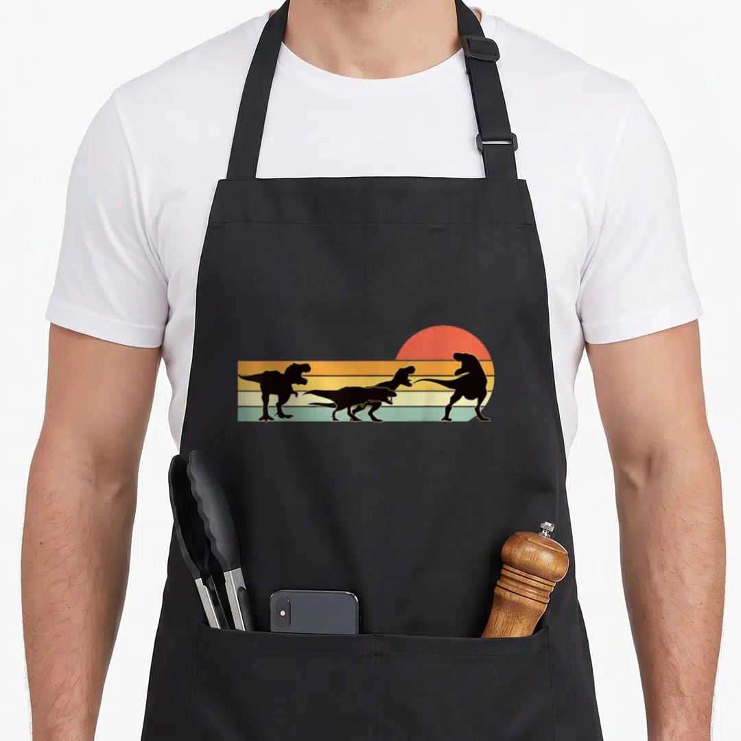 Family t rex apron - Three black dinosaur silhouettes marching across horizontal orange, yellow, and green stripes — shown on a black cotton canvas apron against a white background
