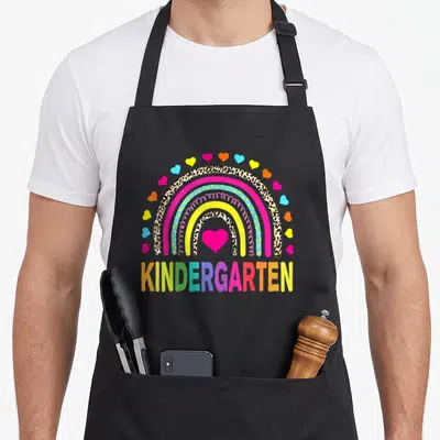 Squad kindergarten teacher apron - Rainbow with leopard print bands, pink heart, and multicolored KINDERGARTEN text — shown on a black cotton canvas apron against a white background