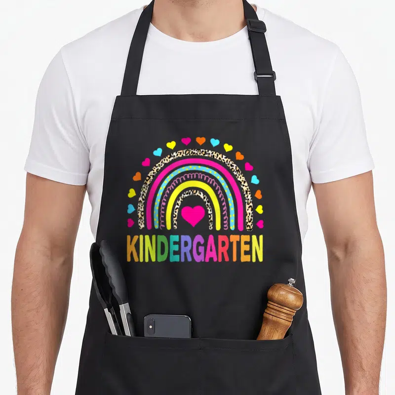 Squad kindergarten teacher apron - Rainbow with leopard print bands, pink heart, and multicolored KINDERGARTEN text — shown on a black cotton canvas apron against a white background