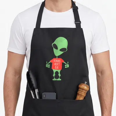 Arent funny alien apron - Green cartoon alien wearing a red shirt with white text HUMANS AREN'T REAL — shown on a black cotton canvas apron against a white background