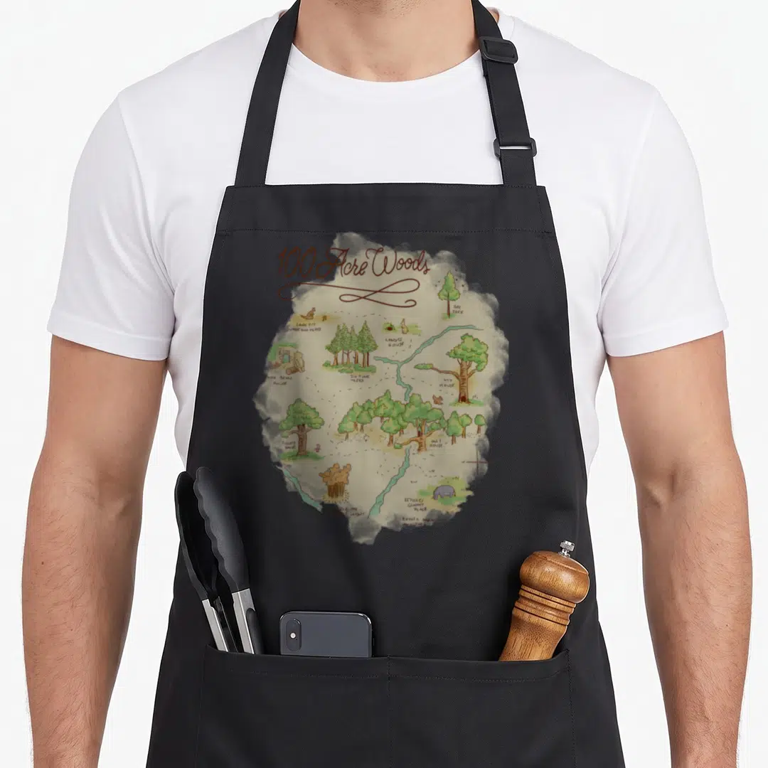 Acre wood apron - Watercolor map of 100 Acre Woods with sandy pit, six pine trees, and character homes — shown on a black cotton canvas apron against a white background