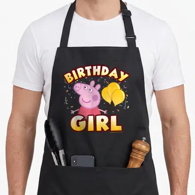 Confetti birthday apron - Pink pig holding three yellow balloons with BIRTHDAY GIRL text and confetti — shown on a black cotton canvas apron against a white background