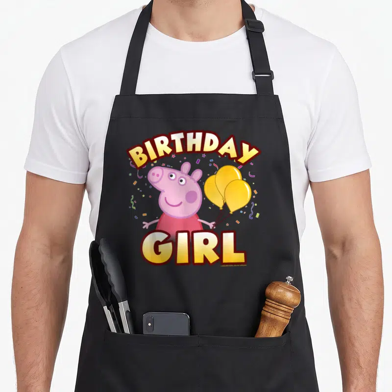 Confetti birthday apron - Pink pig holding three yellow balloons with BIRTHDAY GIRL text and confetti — shown on a black cotton canvas apron against a white background