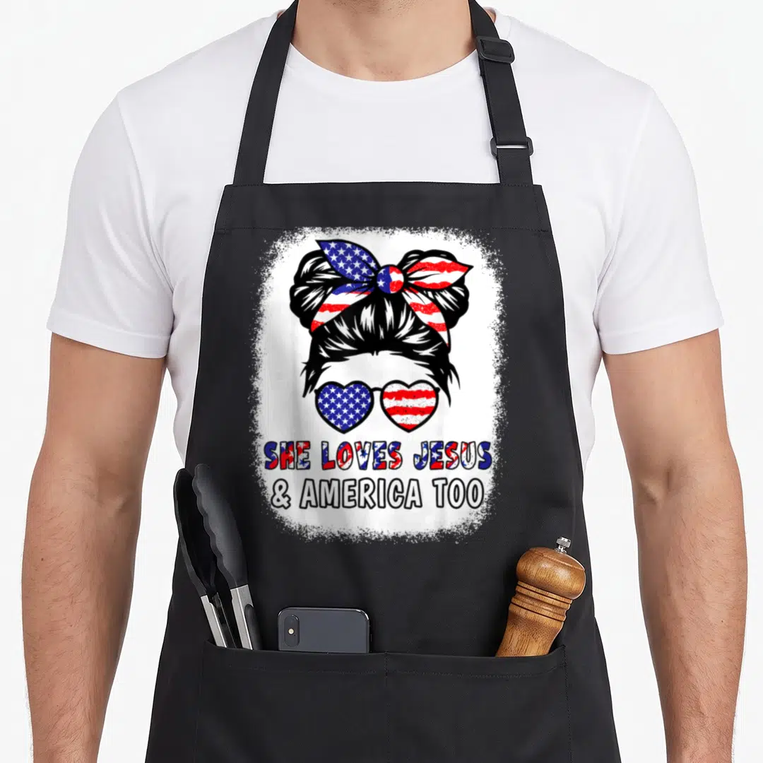 Jesus 4th of july apron - Woman head with bun, flag bow, heart sunglasses, and She Loves Jesus America Too text — shown on a black cotton canvas apron against a white background