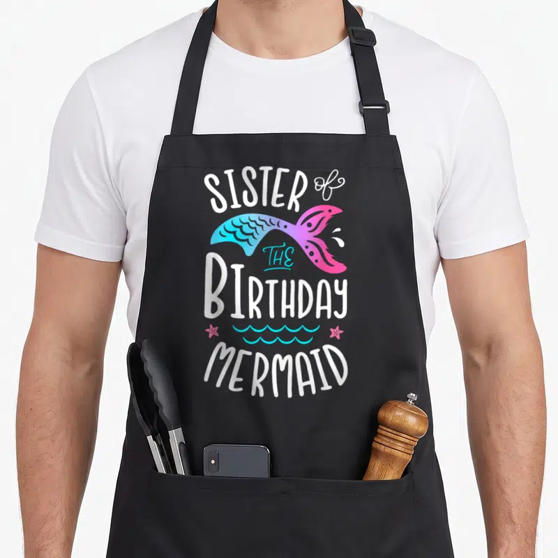 Birthday mermaid family cooking aprons - Sister of the Birthday Mermaid text with cyan pink tail starfish and waves — shown on a black cotton canvas apron against a white background