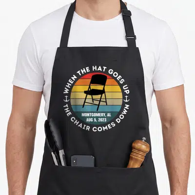 Alabama folding aprons - Black chair on multi-colored striped circle with white text reading WHEN THE HAT GOES UP — shown on a black cotton canvas apron against a white background