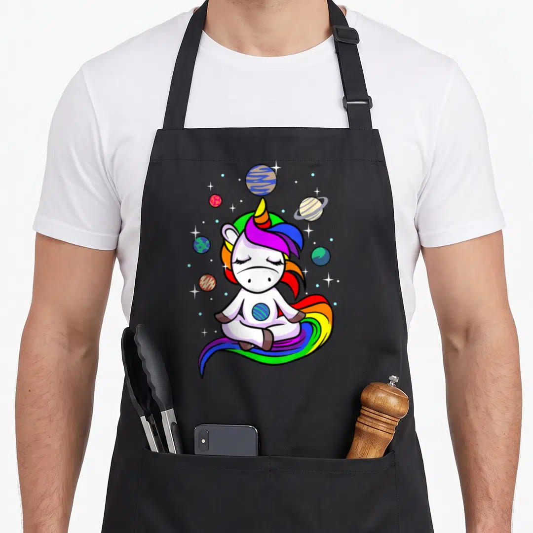 Meditating unicorn apron - White cartoon unicorn meditating among colorful planets and stars with a rainbow tail — shown on a black cotton canvas apron against a white background