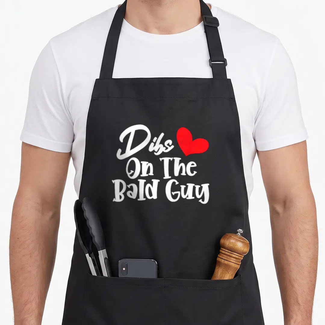 Bald wife apron - White Dibs On The Bald Guy text stacked with a red heart symbol — shown on a black cotton canvas apron against a white background