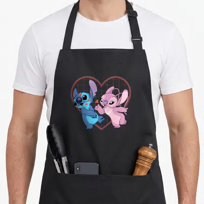 Kiss angels and aprons - Blue and pink cartoon characters in front of a red outlined heart with a grid pattern — shown on a black cotton canvas apron against a white background