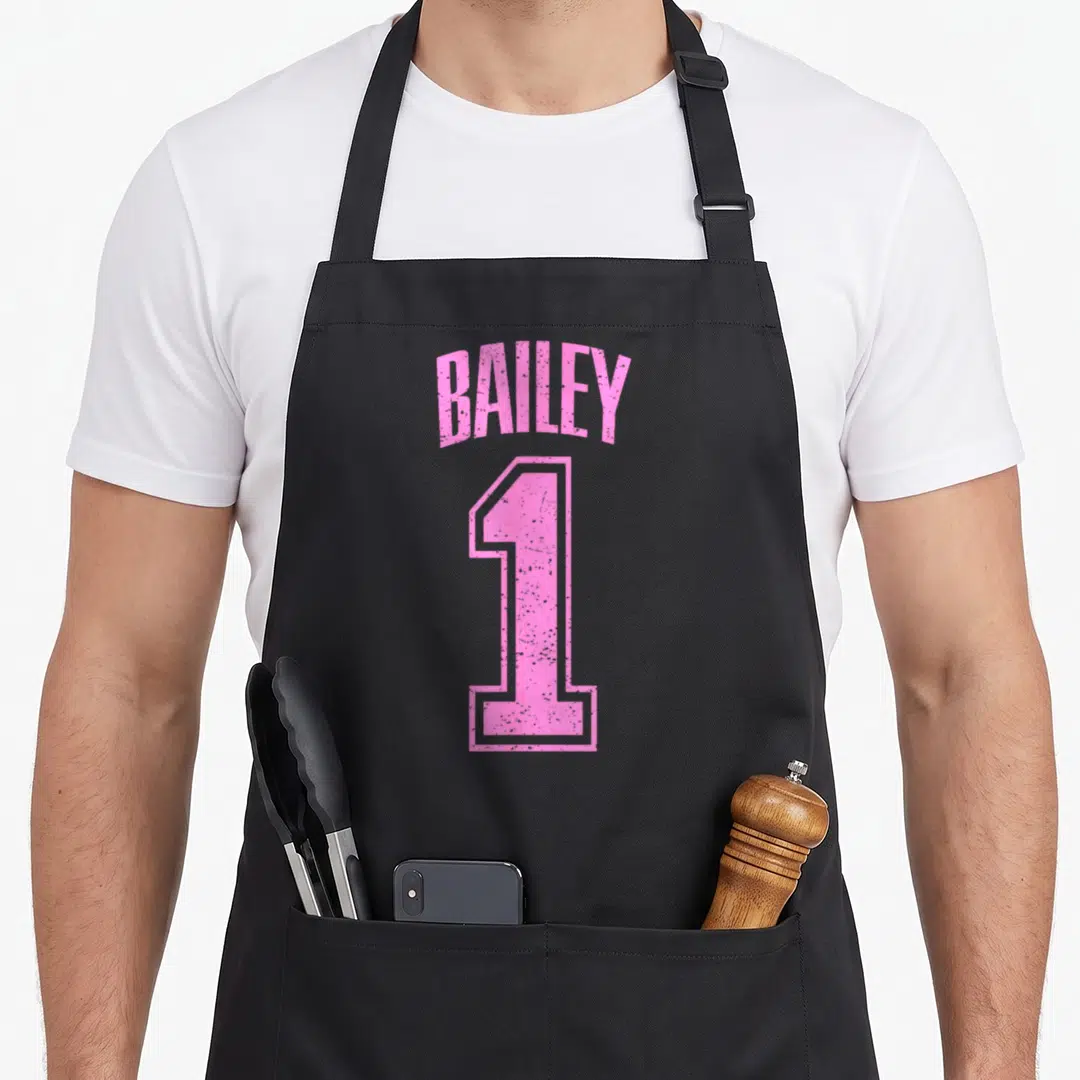 Number baileys apron - Pink arched text reading BAILEY above a large outlined number 1 — shown on a black cotton canvas apron against a white background