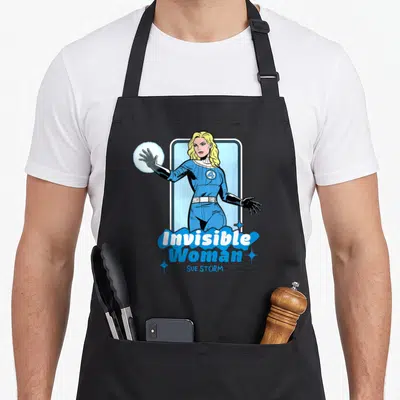 Four invisible first steps apron - Blonde woman in blue suit holding energy sphere with Invisible Woman SUE STORM text — shown on a black cotton canvas apron against a white background