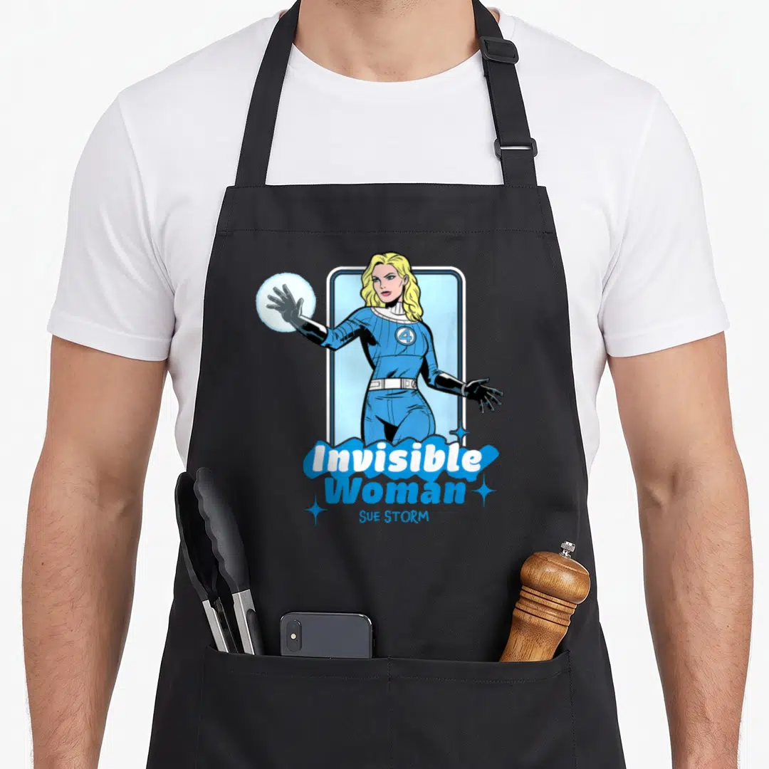 Four invisible first steps apron - Blonde woman in blue suit holding energy sphere with Invisible Woman SUE STORM text — shown on a black cotton canvas apron against a white background