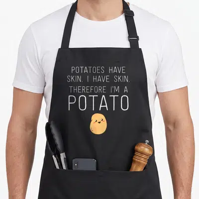 Skin potato apron - White text reading Potatoes Have Skin I Have Skin Therefore I'm Potato above a tan cartoon potato — shown on a black cotton canvas apron against a white background