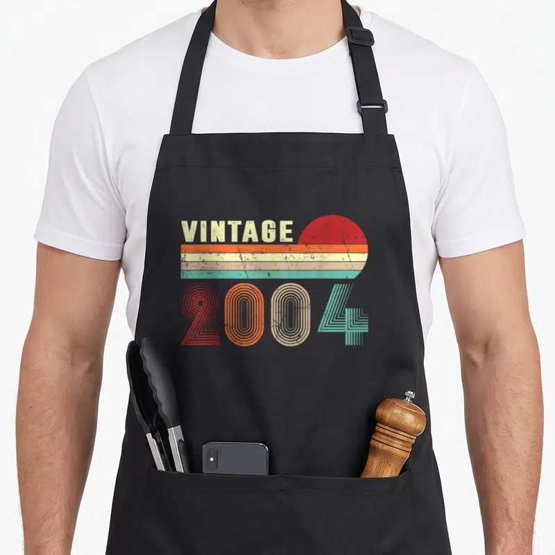 22nd birthday apron - Retro 2004 text design with striped sun and concentric number lines — shown on a black cotton canvas apron against a white background