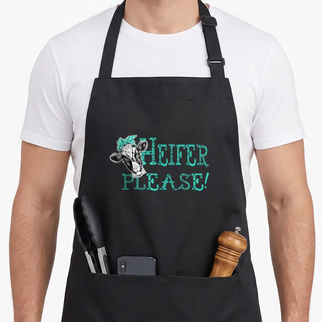 Heifer southern aprons - Cartoon cow head with turquoise bow next to floral text HEIFER PLEASE! — shown on a black cotton canvas apron against a white background