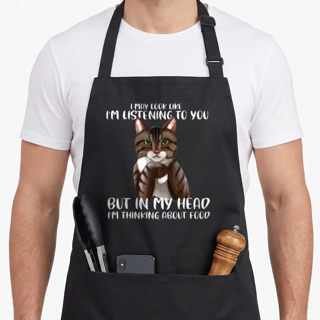 May cat apron - Cartoon brown and white cat with green eyes and food-themed quote text — shown on a black cotton canvas apron against a white background