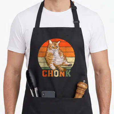 Overweight cat apron - Fat orange cat sitting before striped retro sun with CHONK text below — shown on a black cotton canvas apron against a white background