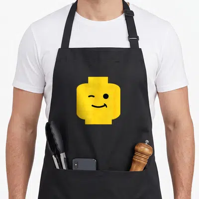 Winking face apron - Yellow toy brick head with winking eye and smiling mouth in minimalist black art style — shown on a black cotton canvas apron against a white background