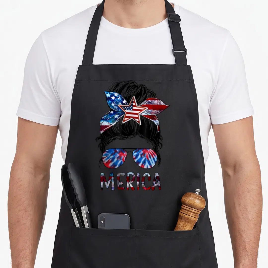 Merica messy aprons - Cartoon woman bun with flag bandana, tie-dye sunglasses, and MERICA text in red white blue — shown on a black cotton canvas apron against a white background