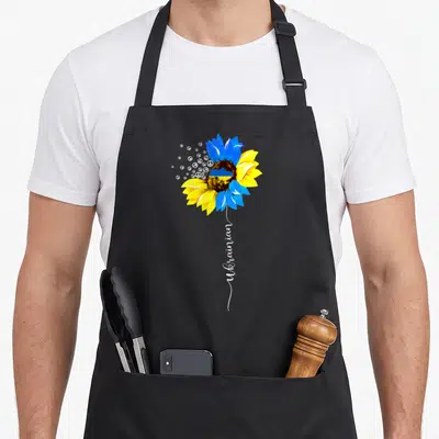 Funny flag ukrainian aprons - Blue and yellow sunflower with peace symbols and vertical Ukrainian text — shown on a black cotton canvas apron against a white background
