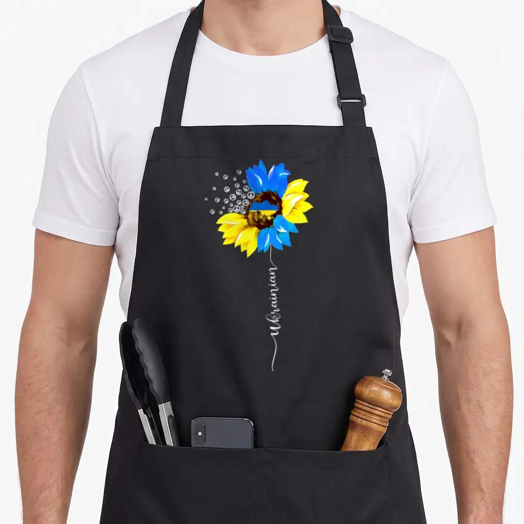 Funny flag ukrainian aprons - Blue and yellow sunflower with peace symbols and vertical Ukrainian text — shown on a black cotton canvas apron against a white background