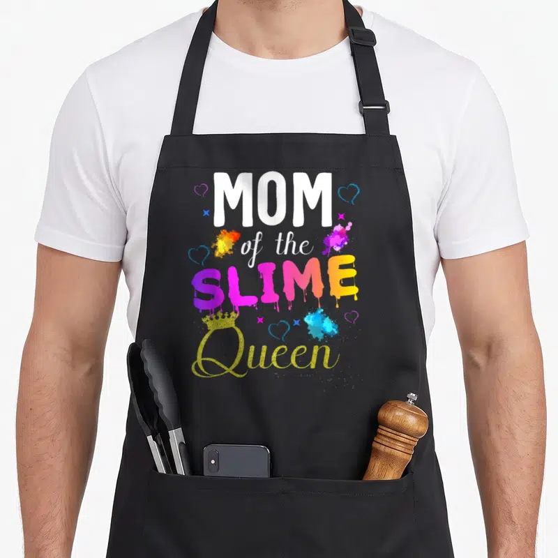 Outfit queen of baking apron - MOM of the SLIME Queen text with dripping font, gold crown, hearts stars paint splatters — shown on a black cotton canvas apron against a white background
