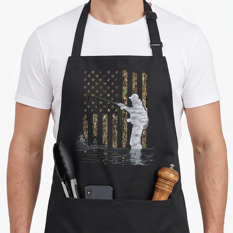 Camouflage fisherman apron - Fly fisherman silhouette casting line over camo flag and water reflection with mountains — shown on a black cotton canvas apron against a white background