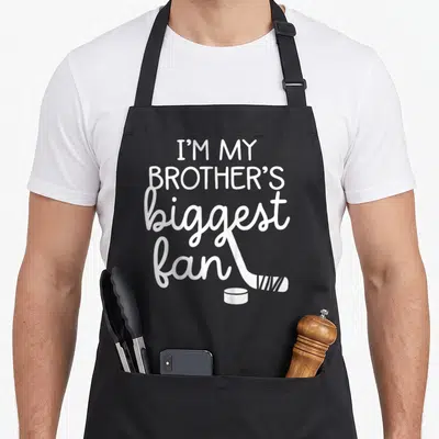 Ice teyliedesign apron - Stacked white text I'M MY BROTHER'S biggest fan with hockey stick and puck — shown on a black cotton canvas apron against a white background