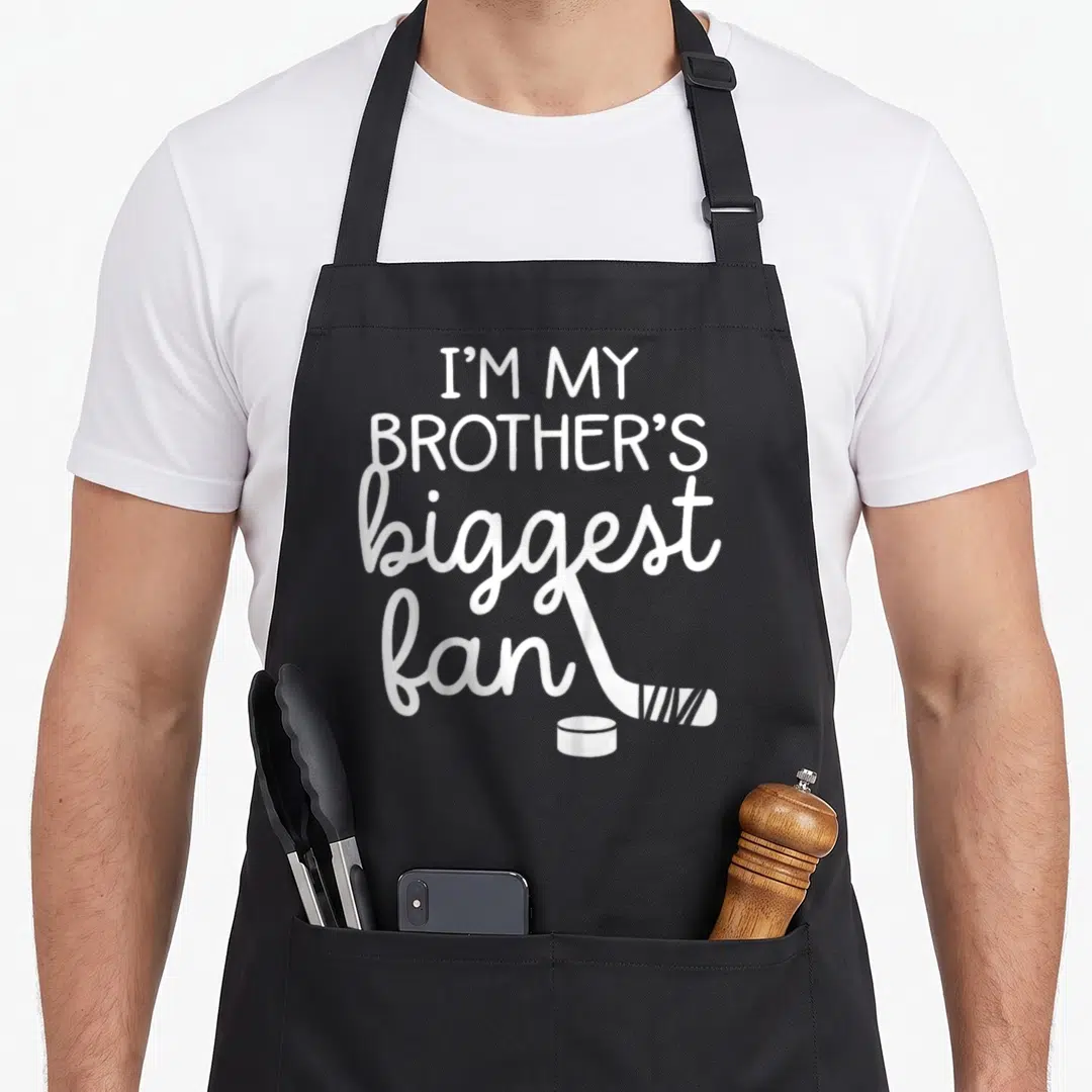 Ice teyliedesign apron - Stacked white text I'M MY BROTHER'S biggest fan with hockey stick and puck — shown on a black cotton canvas apron against a white background