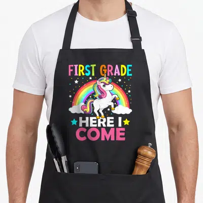 Grade come unicorn apron - Stacked FIRST GRADE HERE I COME text with rearing unicorn and rainbow — shown on a black cotton canvas apron against a white background