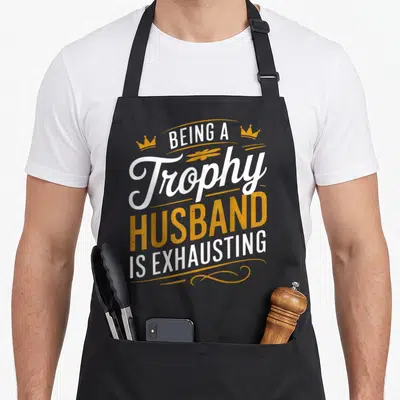Exhausting husband apron - Stacked white and orange text reading Being A Trophy Husband Is Exhausting with two small — shown on a black cotton canvas apron against a white background