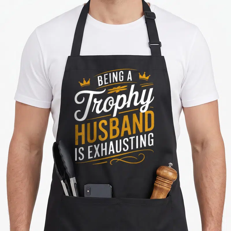 Exhausting husband apron - Stacked white and orange text reading Being A Trophy Husband Is Exhausting with two small — shown on a black cotton canvas apron against a white background