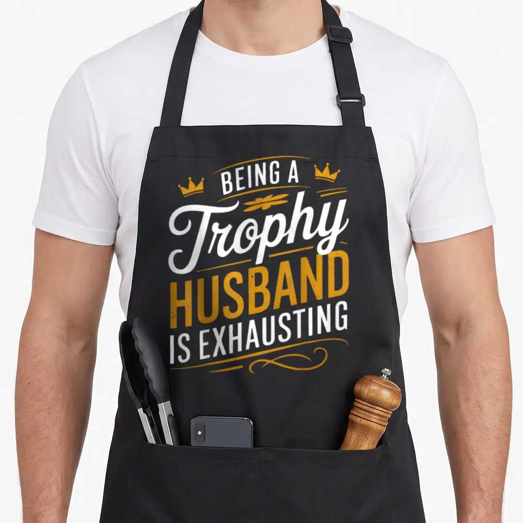 Exhausting husband apron - Stacked white and orange text reading Being A Trophy Husband Is Exhausting with two small — shown on a black cotton canvas apron against a white background