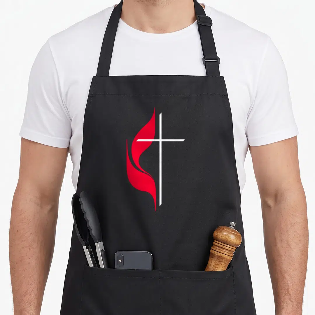 Methodist christian apron - White cross overlapping a red flame in a minimalist style on a black background — shown on a black cotton canvas apron against a white background