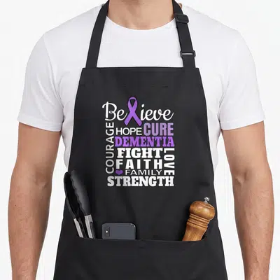 Believe ribbon apron - Purple ribbon replaces X in Believe, surrounded by Dementia, Hope, Courage, Love, Family — shown on a black cotton canvas apron against a white background