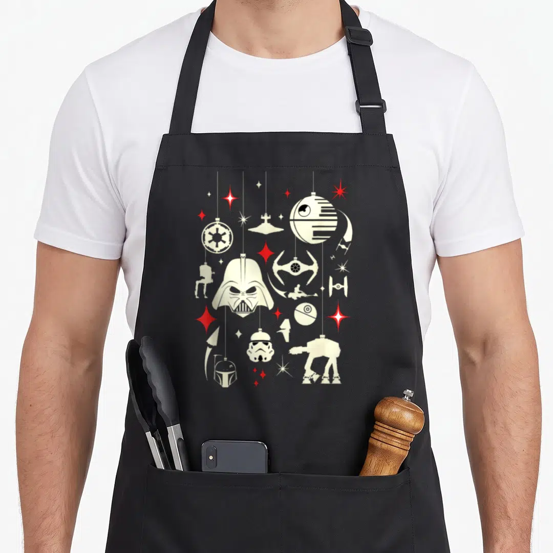Ornament holiday apron - Cream and red Star Wars silhouettes hanging like ornaments with scattered stars on black fabric — shown on a black cotton canvas apron against a white background