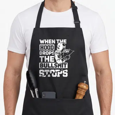 Hood welder apron - White stacked text reading WHEN THE HOOD DROPS THE BULLSHIT STOPS with a welder holding a torch — shown on a black cotton canvas apron against a white background