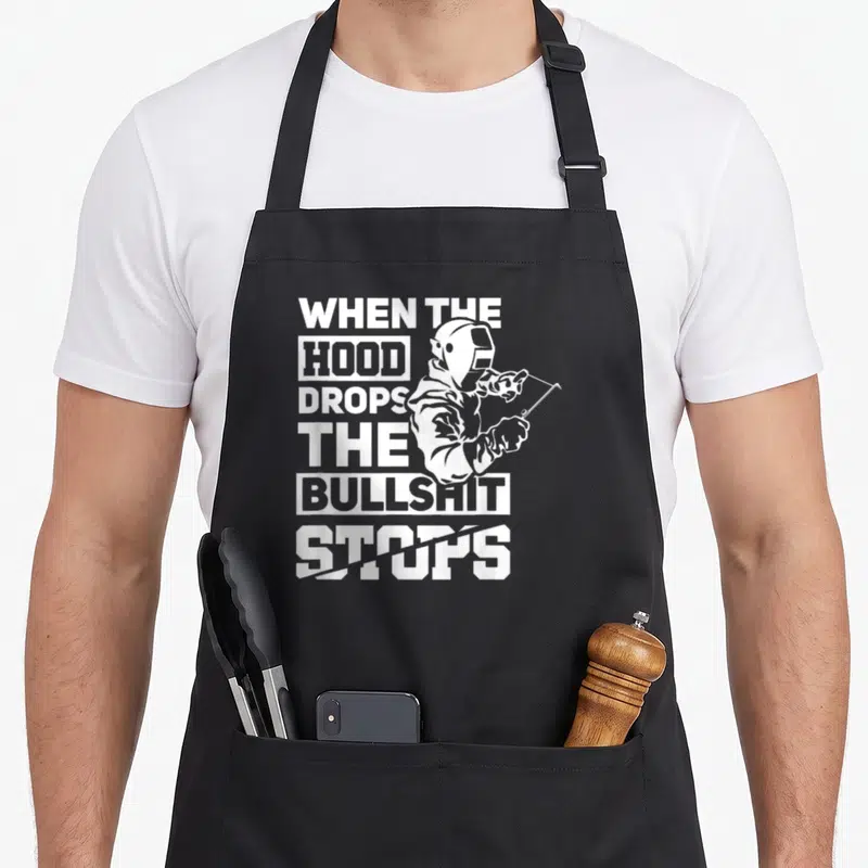 Hood welder apron - White stacked text reading WHEN THE HOOD DROPS THE BULLSHIT STOPS with a welder holding a torch — shown on a black cotton canvas apron against a white background