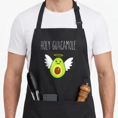 Angel avocado apron - Cartoon avocado with wings and halo under holy guacamole text in white, green and yellow — shown on a black cotton canvas apron against a white background