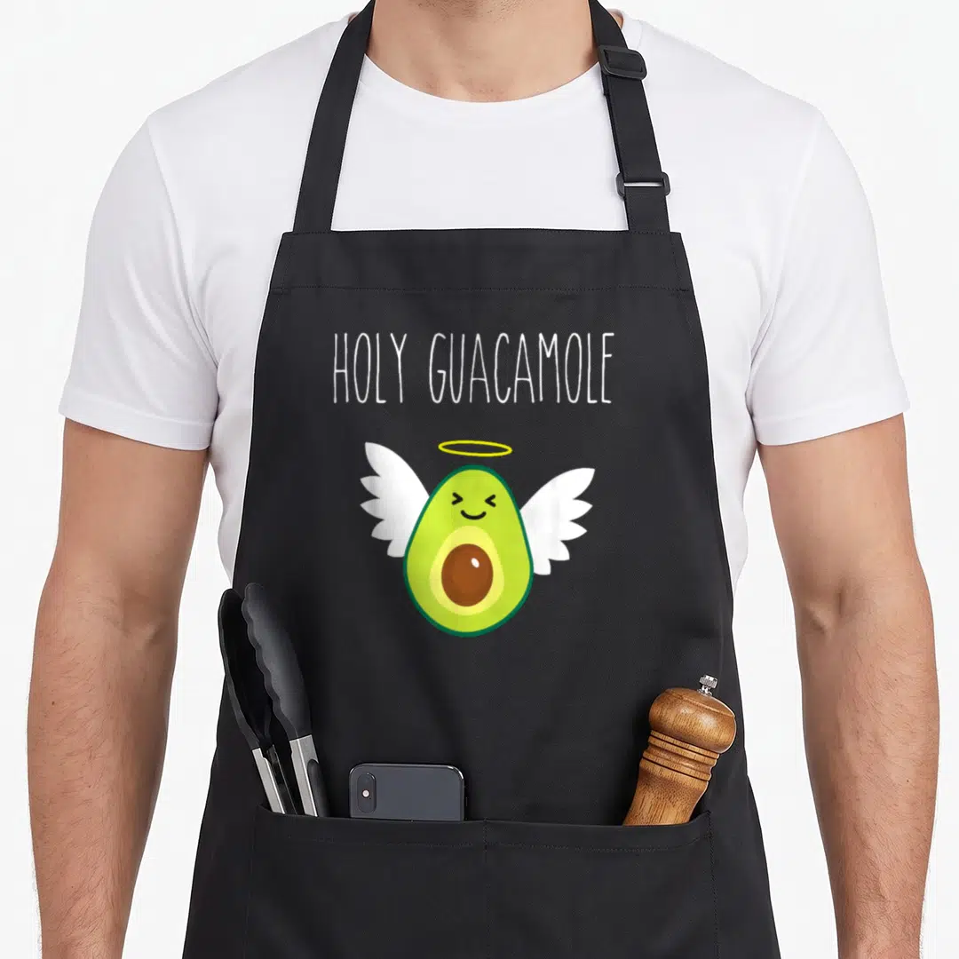 Angel avocado apron - Cartoon avocado with wings and halo under holy guacamole text in white, green and yellow — shown on a black cotton canvas apron against a white background