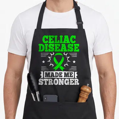 Sprue celiac awareness apron - Green ribbon and text CELIAC DISEASE MADE ME STRONGER with white stars and lines — shown on a black cotton canvas apron against a white background