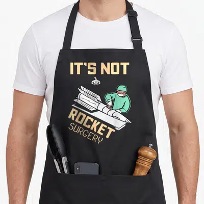 Operating surgeon apron - Cartoon surgeon in green scrubs operating on a white rocket under a lamp with peach text IT'S — shown on a black cotton canvas apron against a white background