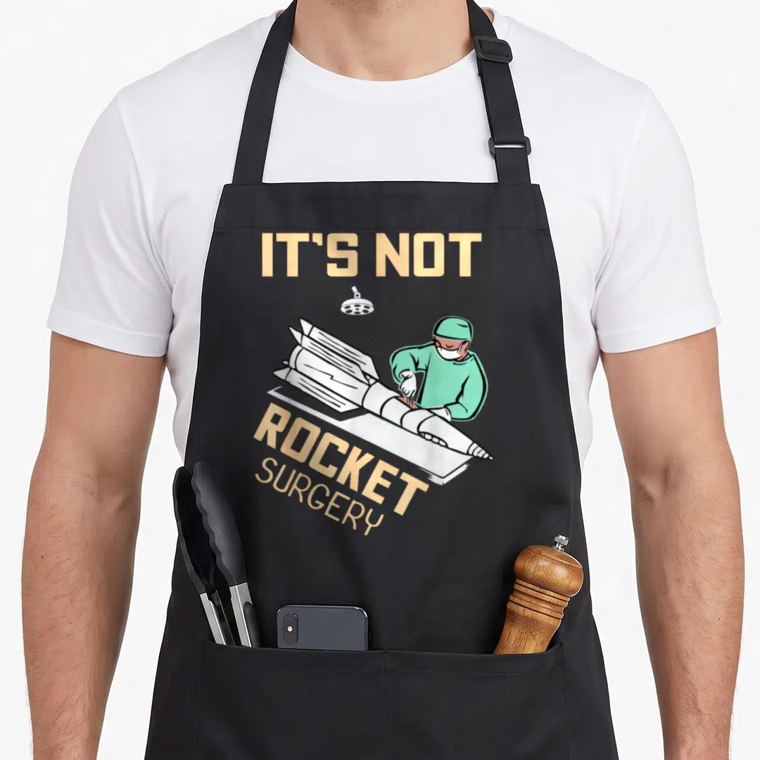 Operating surgeon apron - Cartoon surgeon in green scrubs operating on a white rocket under a lamp with peach text IT'S — shown on a black cotton canvas apron against a white background