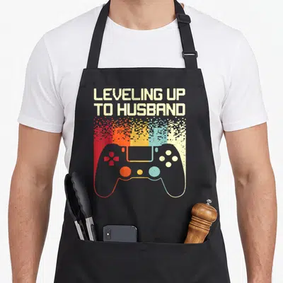 Future groom husband apron - Retro game controller with white red orange yellow blue pixels and leveling up to husband — shown on a black cotton canvas apron against a white background